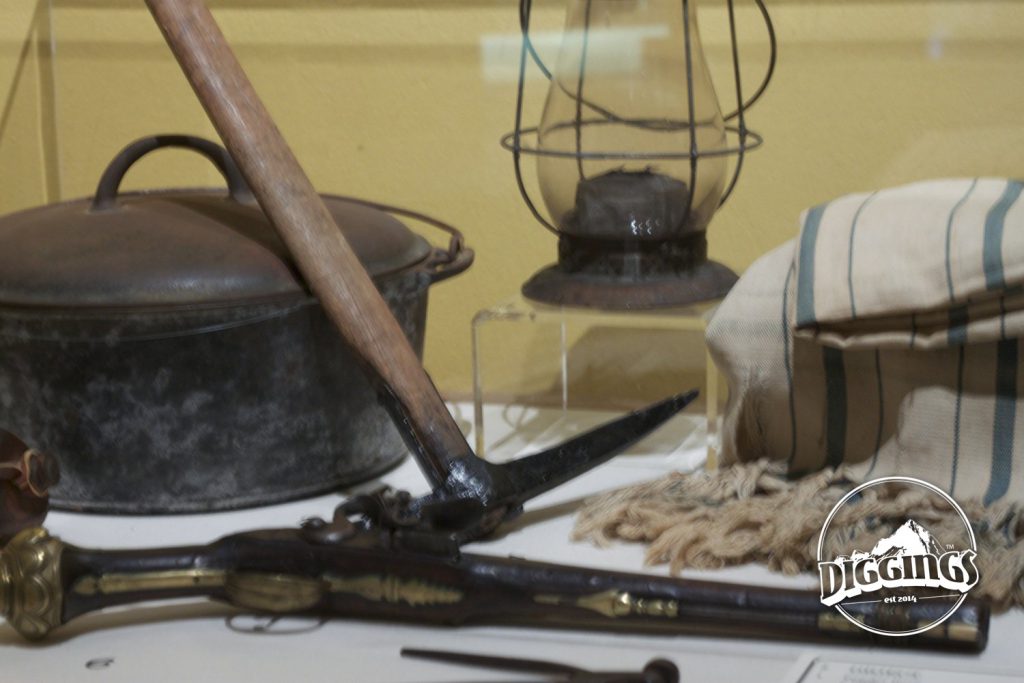 Display of common tools that gold miners would have had during the California Gold Rush, including: pick, dutch oven, lamp, blanket, and pistol.